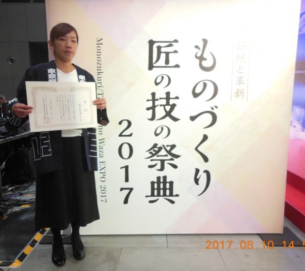 ものづくり匠の技の祭典2017の看板の前で表彰状を持つ福吉奈津子