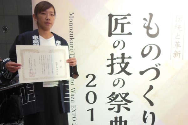 ものづくり匠の技の祭典2017の看板の前で表彰状を持つ福吉奈津子