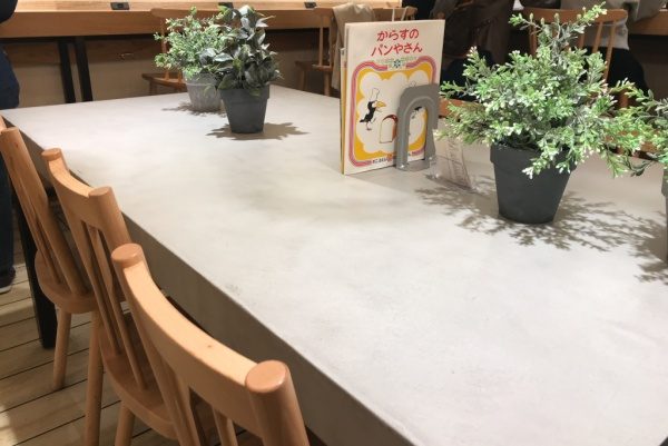 パン屋さんのモールテックスグレーのテーブル、机の上には観葉植物や本が置いてある、周りに椅子もある、絵本がある