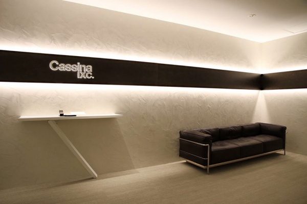 白いオルトレマテリアで施工された壁、カッシーナ・イクスシー青山オフィスエントランス、テーブルがあり壁には社名ロゴ、黒いソファがある